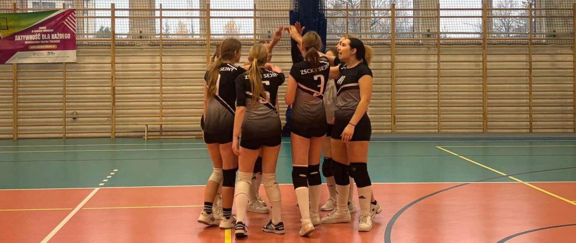 Zawodniczki drużyny siatkarskiej stoją razem w kręgu, trzymając ręce w górze w geście mobilizacji.
