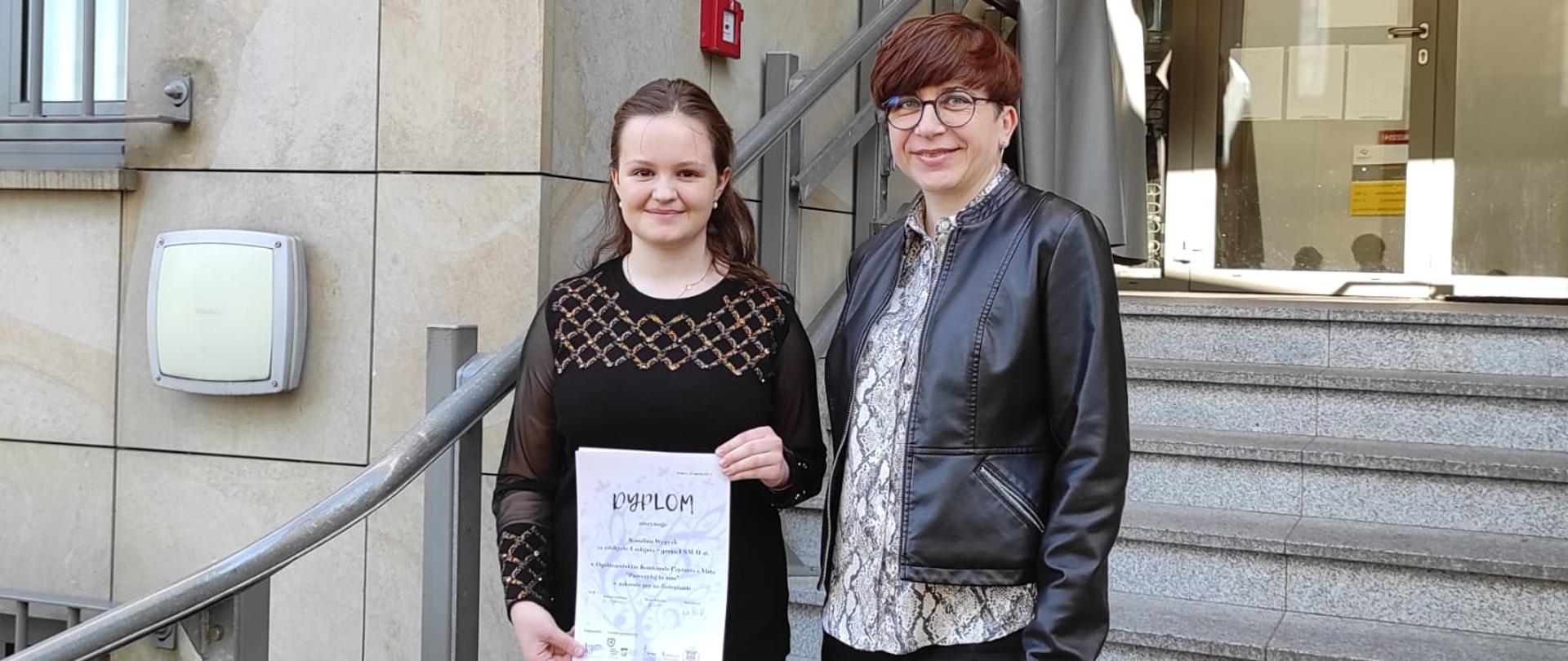 Kolorowe zdjęcie na schodach przed szkołą muzyczną w Krakowie Karoliny Wypych i jej nauczycielki Pani Katarzyny Leśnik. Obie Panie stoją obok siebie a uczennica trzyma rękach dyplom za zajęcie pierwszego miejsca.