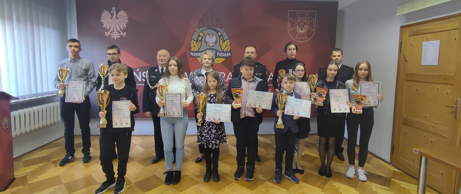 Zdjęcie przedstawia salę konferencyjną Komendy Powiatowej PSP w Łańcucie. Na tle logo PSP stoją wszyscy uczestnicy konkursu oraz wicestarosta łańcucki Pani Barbara Pilawa-Kraus, prezes zarządu powiatowego ZOSP RP w Łańcucie dh Adam Rajzer, zastępca komendanta powiatowego Państwowej Straży Pożarnej w Łańcucie st. bryg. Jacek Rejman przewodniczący komisji kpt. Dariusz Kaczor