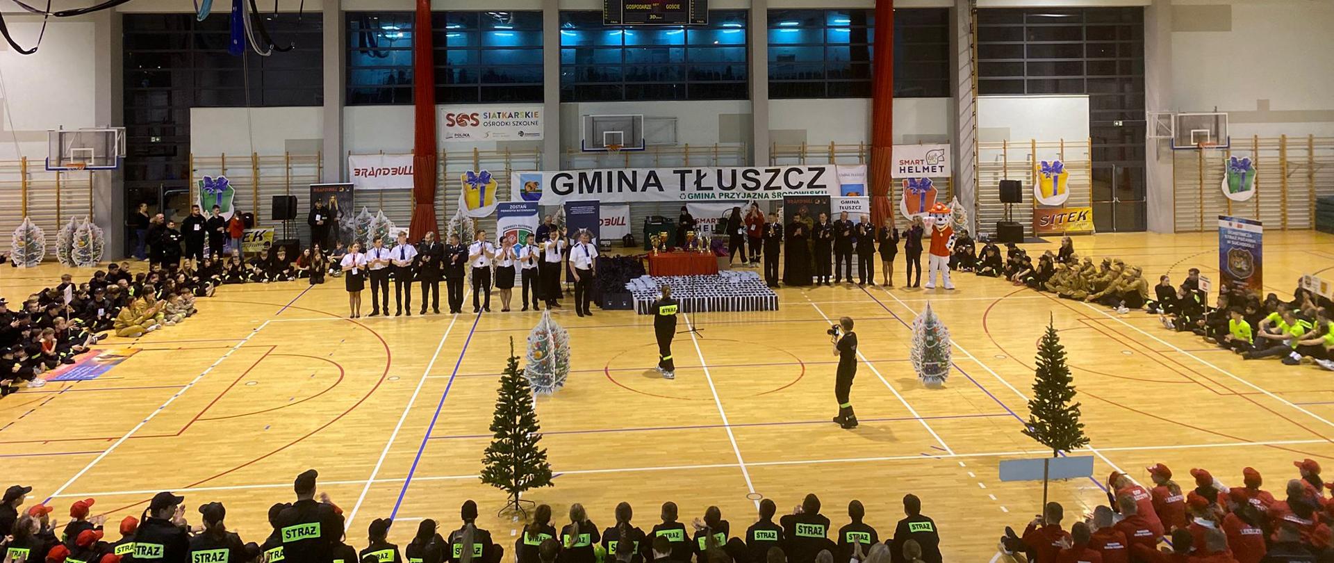 Szerokie ujęcie całej hali sportowej podczas uroczystości. Po obu stronach parkietu siedzą grupy młodych strażaków w jednolitych strojach. W centrum ustawieni są prowadzący i goście wydarzenia, a za nimi liczne nagrody, banery i dekoracje, w tym małe sztuczne choinki.