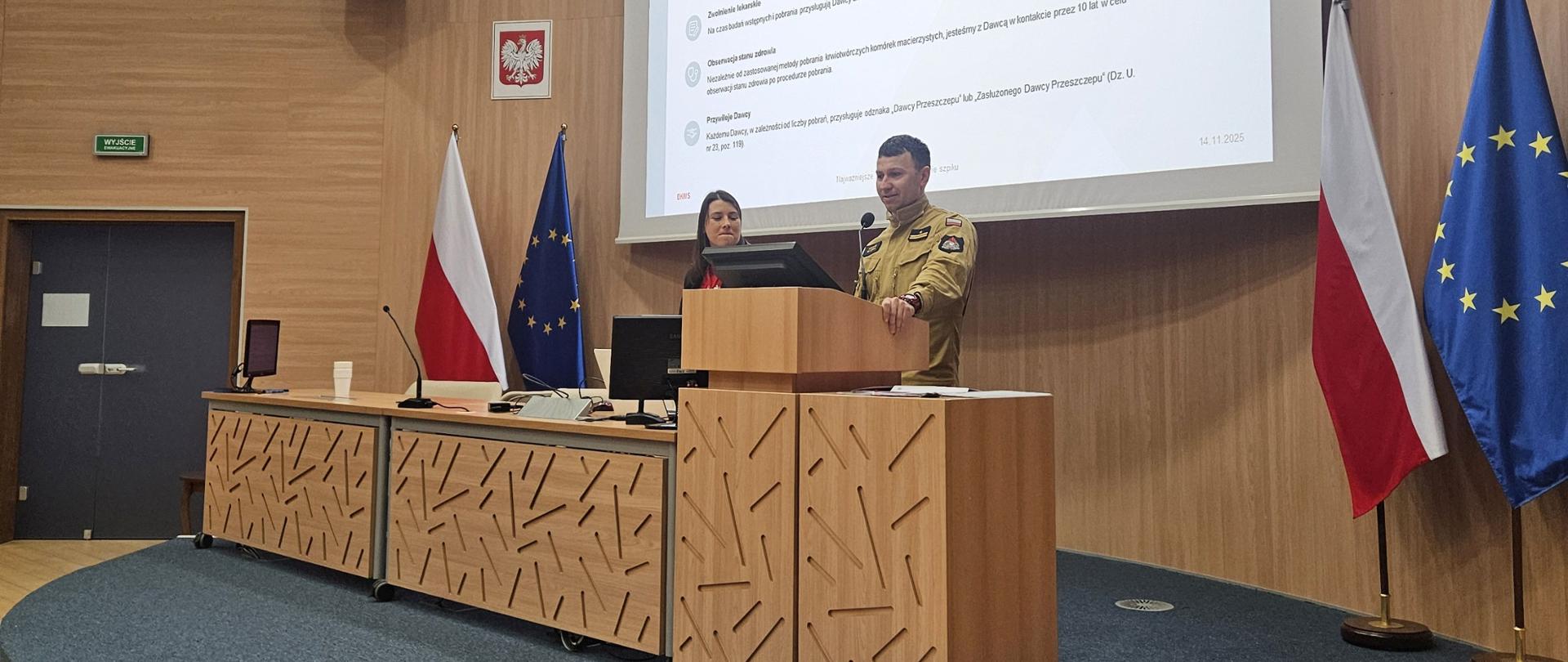 Na zdjęciu widoczna jest aula wykładowa w Centralnej Szkole PSP w Częstochowie. Na podwyższeniu stoi dwóch prelegentów – kobieta i mężczyzna w mundurze strażackim – prowadzących prezentację przy mównicy. Za nimi znajduje się duży ekran z wyświetlonym slajdem dotyczącym informacji dla dawców. Po bokach ustawione są flagi Polski i Unii Europejskiej, a na ścianie widoczny jest krzyż
