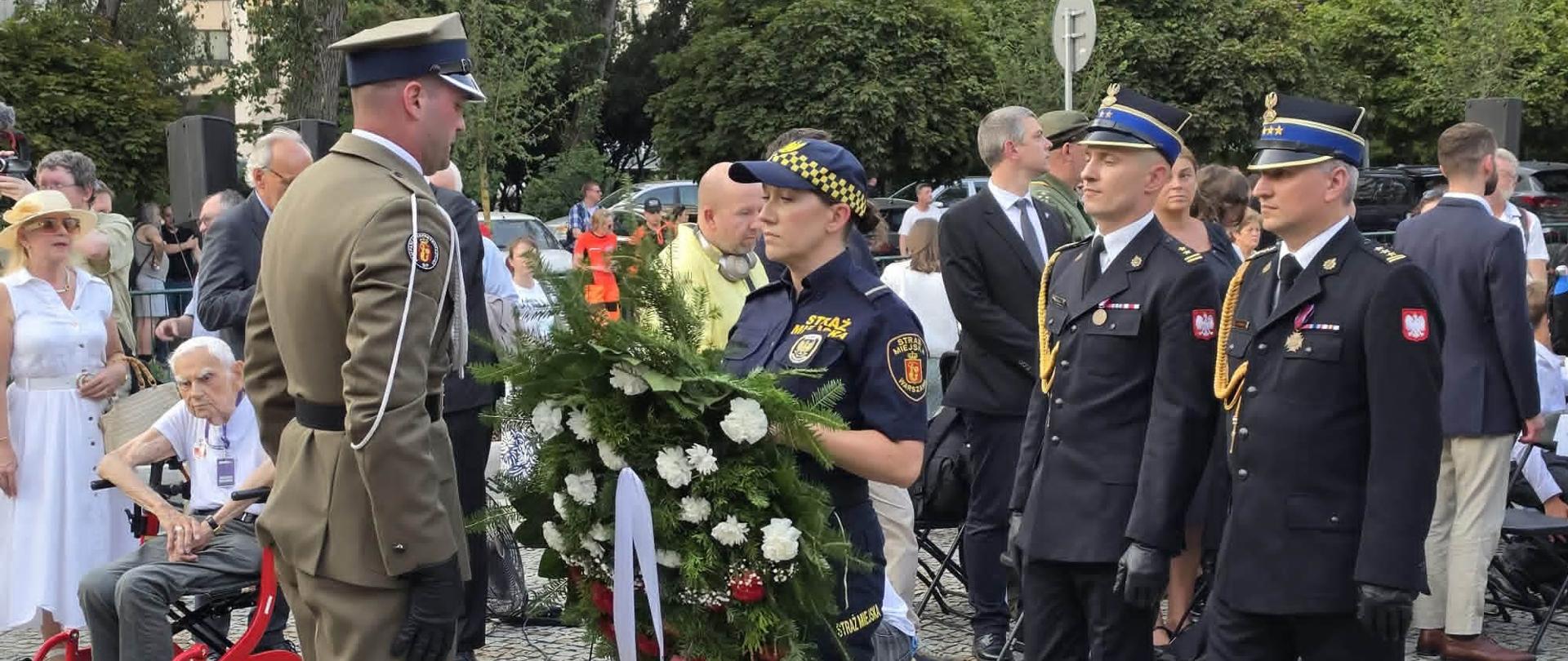 Uroczysta ceremonia składania wieńca na placu wyłożonym kostką brukową. Na pierwszym planie umundurowani funkcjonariusze: żołnierz w beżowym mundurze stoi przodem do strażniczki miejskiej z Warszawy, która trzyma wieniec ze wstęgą. Obok niej maszerują dwaj strażacy w czarnych mundurach galowych. W tle zgromadzeni cywile i zieleń miejska.