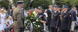 Uroczysta ceremonia składania wieńca na placu wyłożonym kostką brukową. Na pierwszym planie umundurowani funkcjonariusze: żołnierz w beżowym mundurze stoi przodem do strażniczki miejskiej z Warszawy, która trzyma wieniec ze wstęgą. Obok niej maszerują dwaj strażacy w czarnych mundurach galowych. W tle zgromadzeni cywile i zieleń miejska.