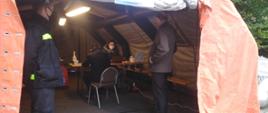 Na zdjęciu namiot koloru pomarańczowego, w namiocie stoi strażak w ubraniu koszarowym obok osoby czekające na szczepienie.