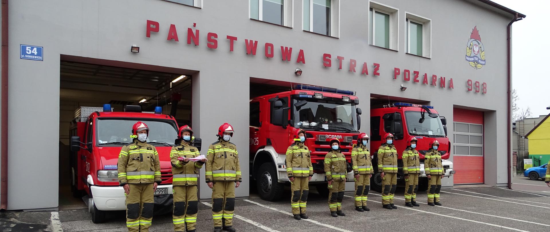 Zdjęcie wykonane przed Komendą Państwowej Straży Pożarnej w Sępólnie Krajeńskim podczas uroczystego podniesienia flagi Państwowej. Strażacy Państwowej Straży Pożarnej w ubraniach specjalnych stoją przed garażami w szeregu na baczność. W tle otwarte garaże oraz trzy samochody gaśnicze.