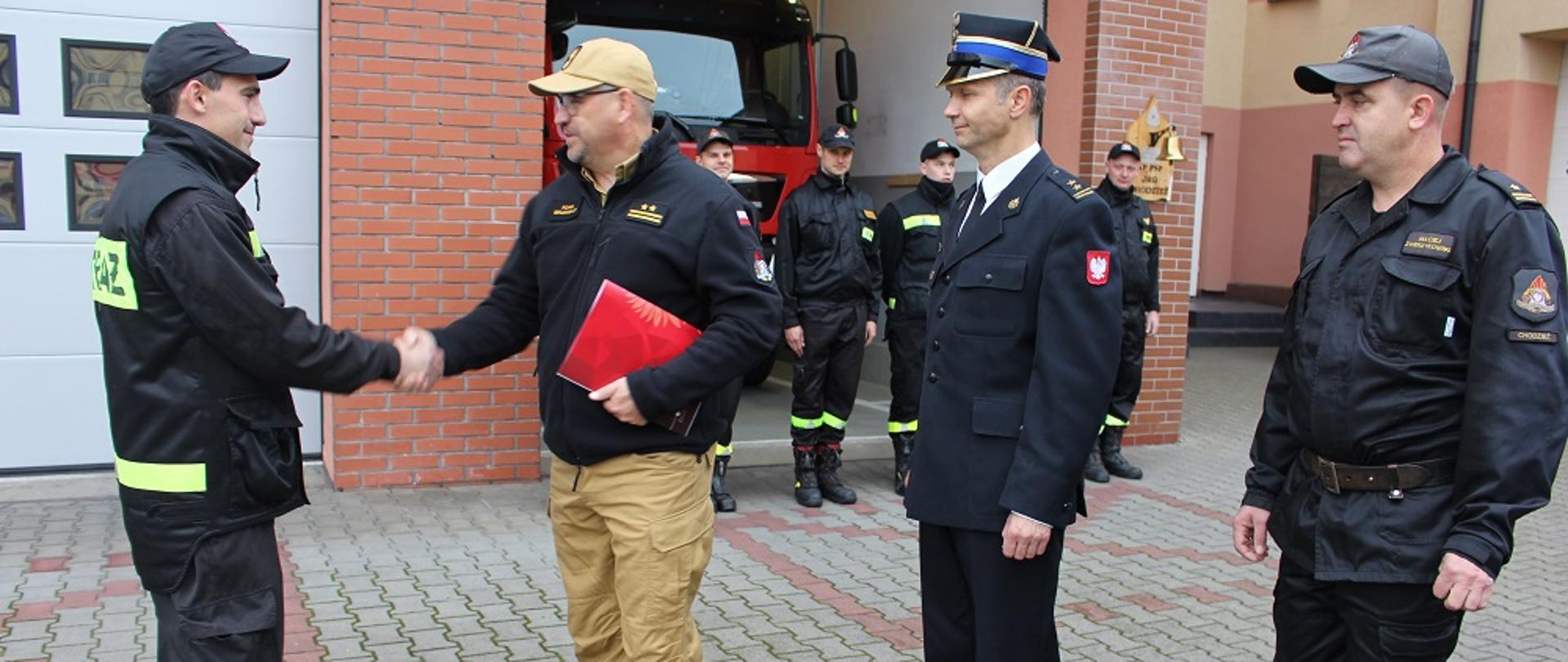 Zdjęcie przedstawia strażaków podczas uroczystej zmiany służby.
W tle budynki.
