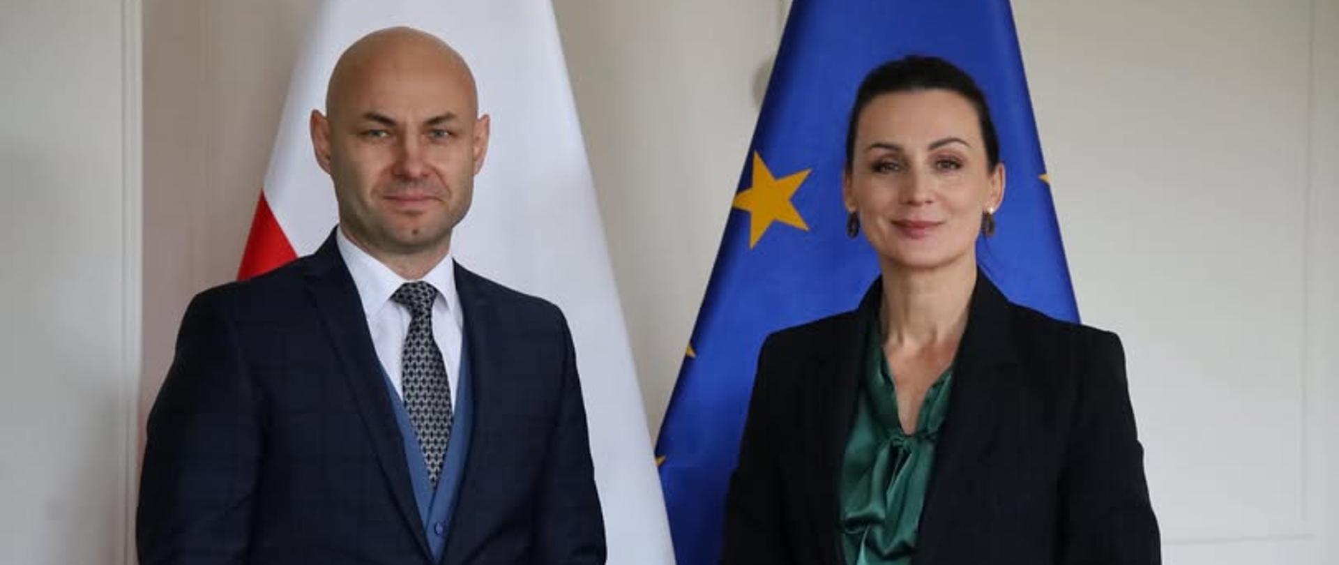 Oficjalne zdjęcie przedstawiające dwoje ludzi - mężczyznę w ciemnym garniturze oraz kobietę w czarnej marynarce i zielonej bluzce. Postacie stoją obok siebie, uśmiechając się do aparatu. W tle widoczne są dwie flagi: Polski oraz Unii Europejskiej.