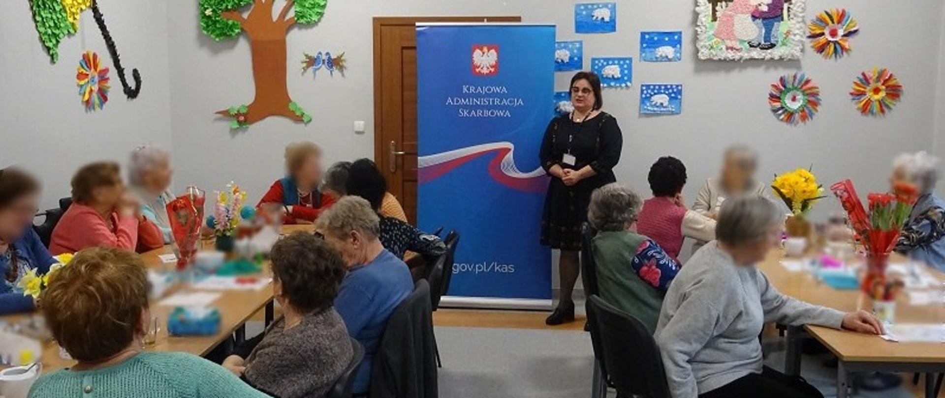 Uczestnicy szkolenia siedzą w sali. Przed nimi stoi ekspertka. Obok niej stoi baner KAS.