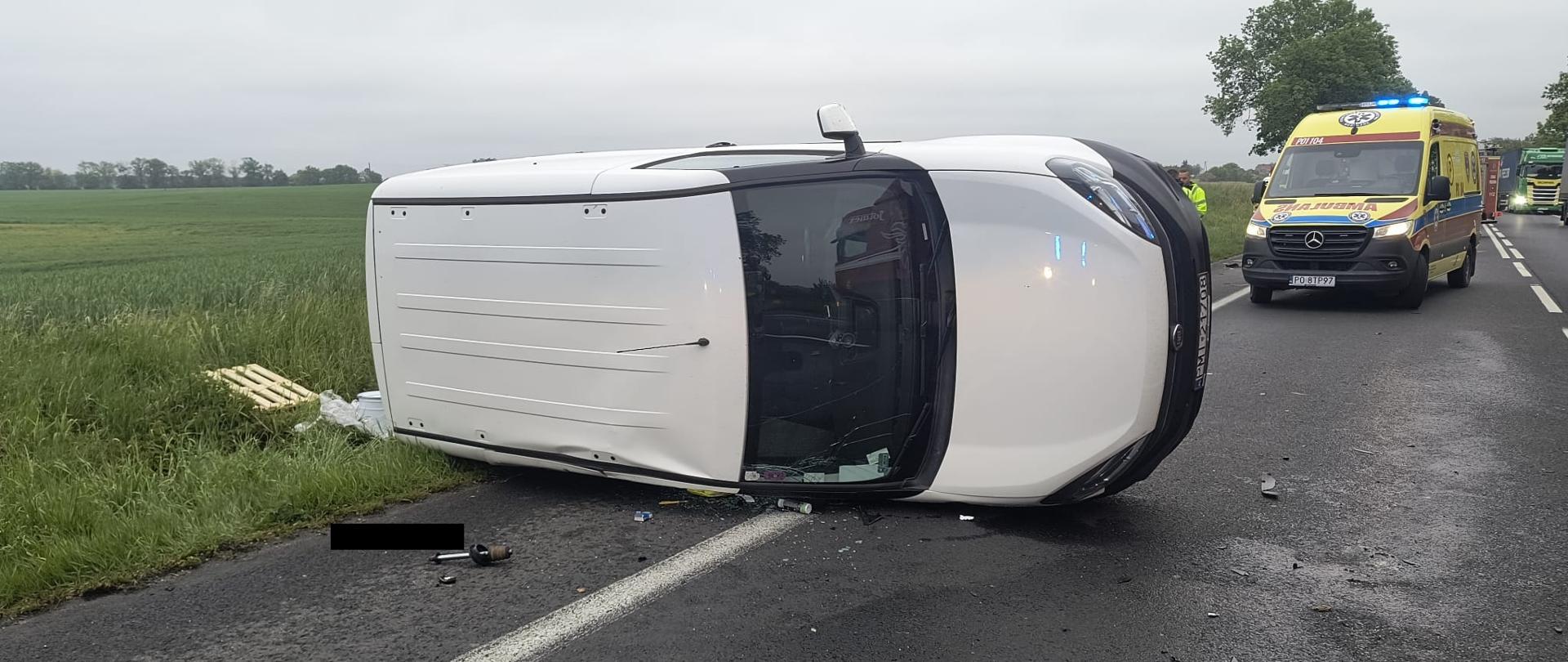 Biały samochód na prawym boku na drodze, za nim karetka pogotowia