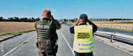 Funkcjonariusz Straży Leśnej i inspektor transportu drogowego stoją przy drodze i obserwują nadjeżdżające pojazdy.