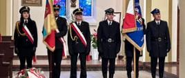 Zdjęcie ukazuje uroczystą chwilę wewnątrz kościoła, podczas Mszy Świętej. W centrum kadru, w pobliżu prezbiterium, stoi sześć osób w galowych mundurach – trzech strażaków i troje strażników miejskich. Dwie osoby trzymają sztandary. Po lewej stronie widoczny jest bukiet kwiatów w barwach narodowych, co sugeruje upamiętnienie ważnego wydarzenia. W tle dominują elementy wyposażenia kościoła, takie jak duży krucyfiks na ścianie i witraże za nim.
Całość oświetlona jest jasnym, ciepłym światłem, które podkreśla powagę i uświęcony charakter miejsca. Kolorystyka jest stonowana, z akcentami bieli i czerwieni na szarfach i sztandarach.
