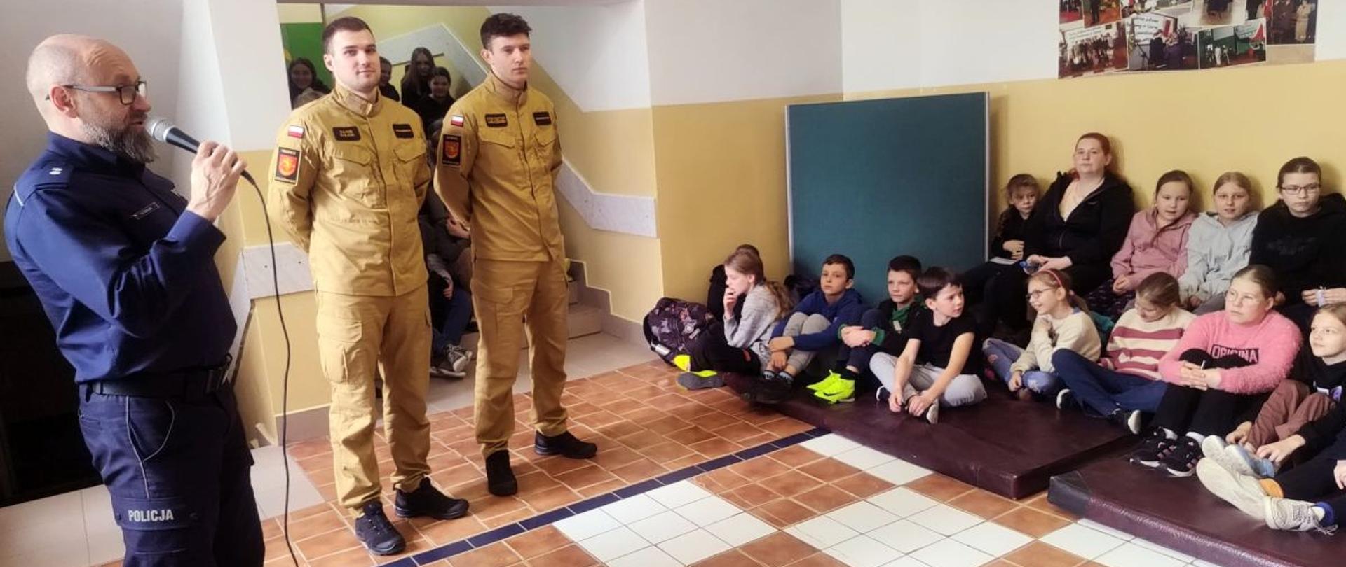 Na zdjęciu widać dwóch strażaków, policjanta, panią nauczycielkę i dzieci które siedzą pod ścianą na materacach.