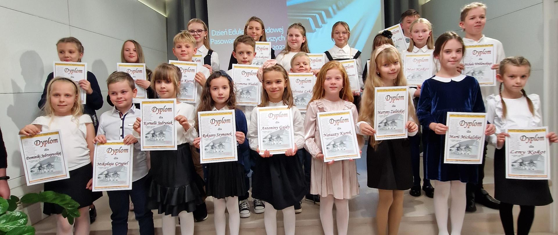 Na zdjęciu widać dzieci stojące w kilku rzędach na stopniach sceny, ubrane na galowo i trzymające w dłoniach dyplomy zwrócone przodem do publiczności. W tle znajduje się wysunięty ekran, na którym wyświetlony jest slajd z napisem Dzień Edukacji Narodowej i klawiaturą fortepianu. 