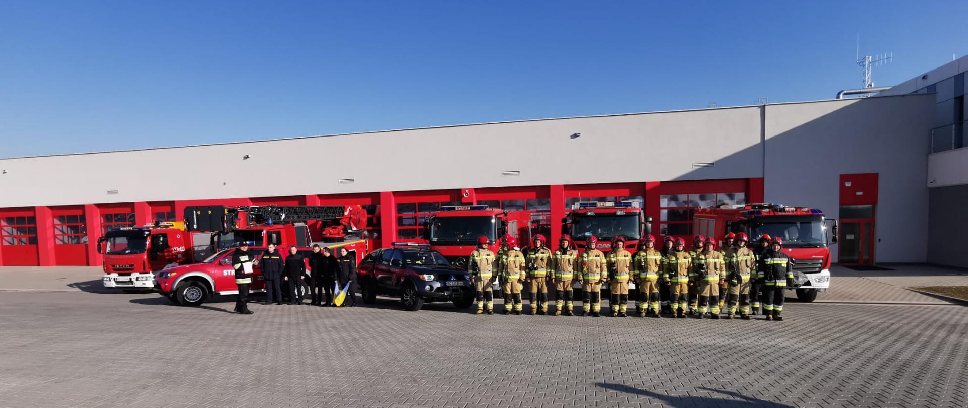 Zdjęcie przedstawia rzeszowskich strażaków w umundurowaniu bojowym stojących na placu zewnętrznym w dwuszeregu przed czterema pojazdami pożarniczymi wystawionymi przed bramami garażu jednostki.