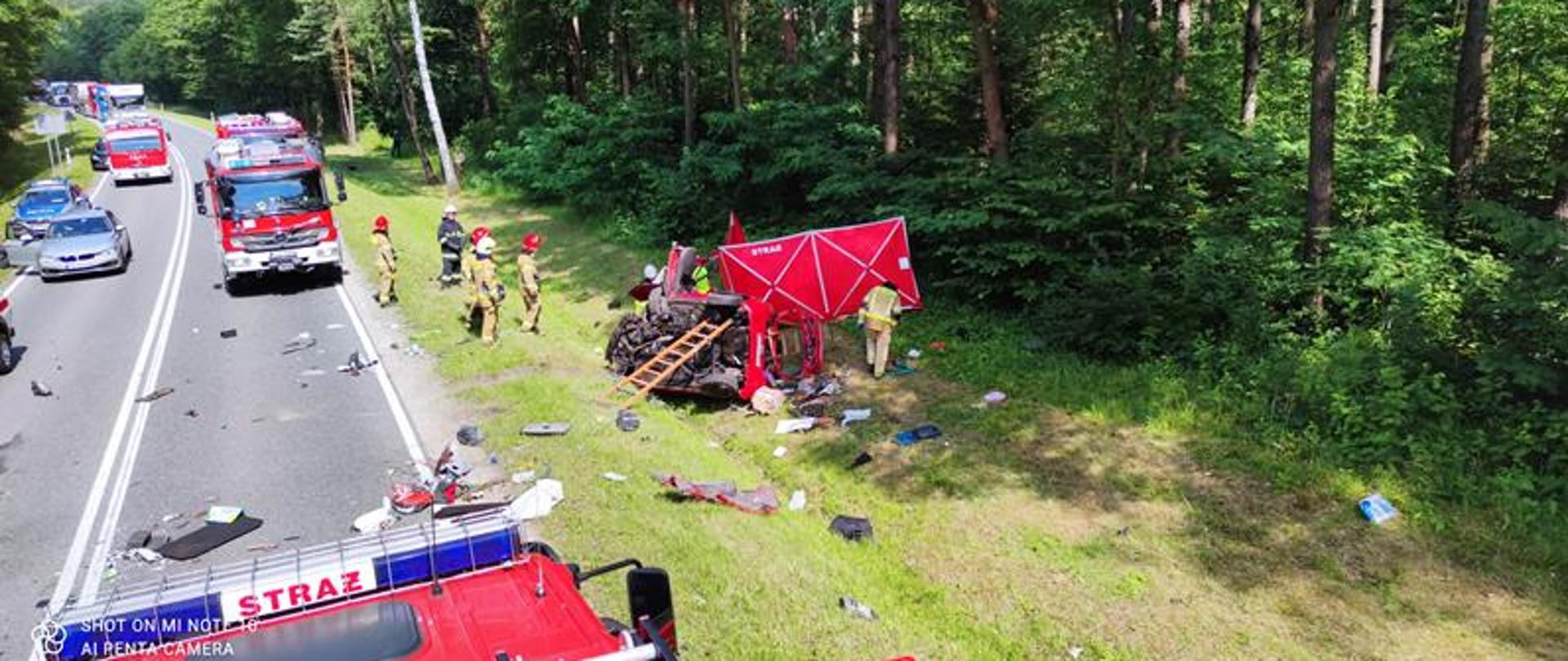 Strażacy ubrani w ubrania specjalne stoją obok rozbitego samochodu osobowego samochodu który znajduje się w rowie. Obok samochodu rozstawiony jest parawan strażacki. Na drodze asfaltowej ustawione są pojazdy pożarnicze.