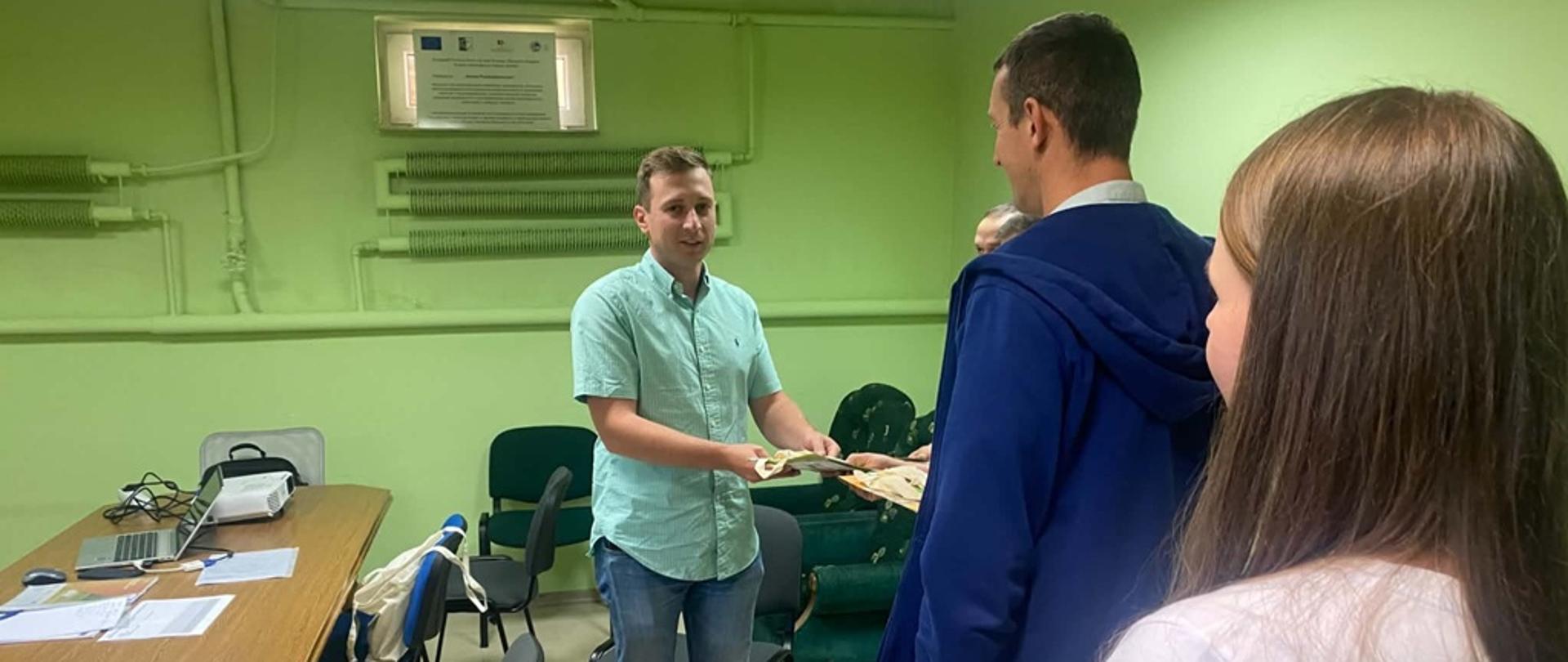Na pierwszym planie widoczny jest mężczyzna ubrany w zielona koszulę wręczający materiały promocyjne. Po prawej stronie widocznych jest dwóch mężczyzn oraz kobieta jeden z mężczyzn ubrany w granatową bluzę, natomiast kobieta ubrana jest w białą koszulkę z krótkim rękawem. Po lewej stronie na brązowym stole widoczny jest szary laptop z rzutnikiem oraz białe kartki z materiałami dotyczącymi szkolenia.