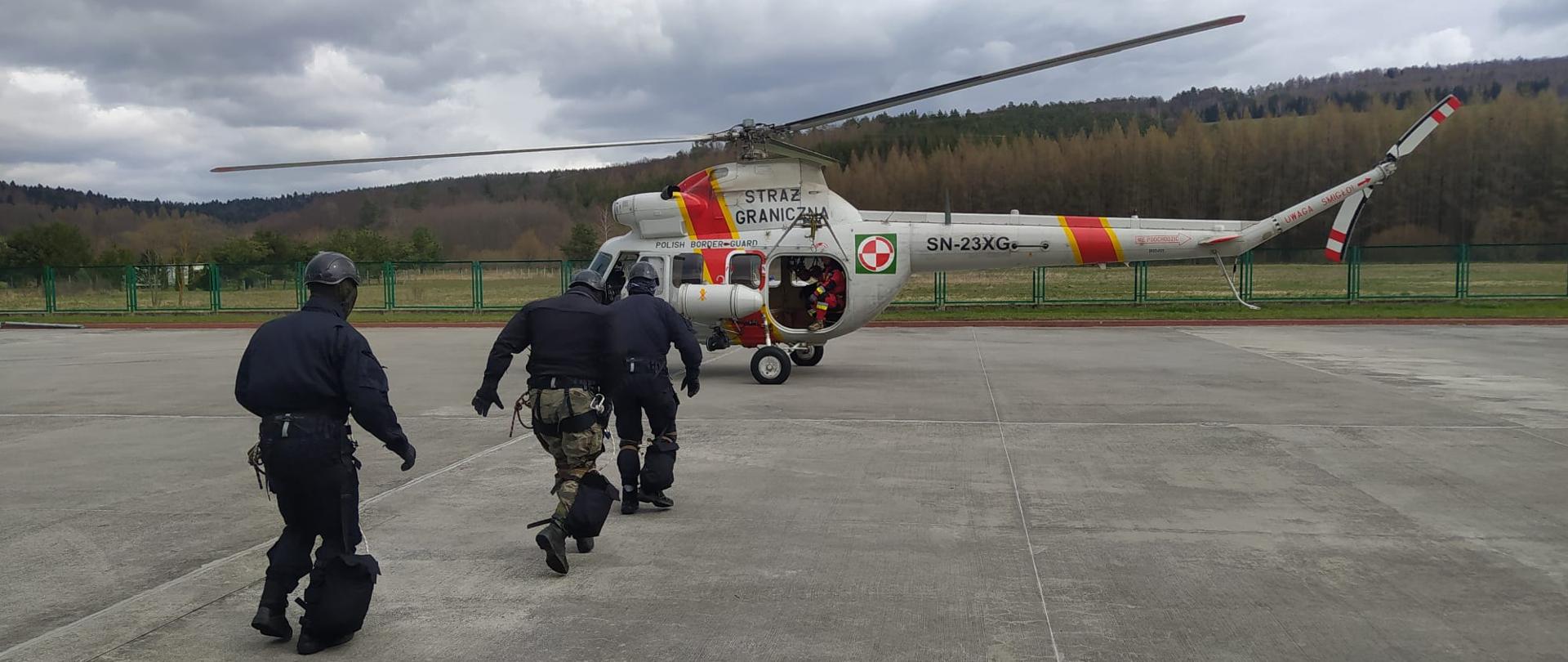 Śmigłowiec w trakcie przyziemienia na płycie lotniska. W jego kierunku biegnie trzech mężczyzn. 