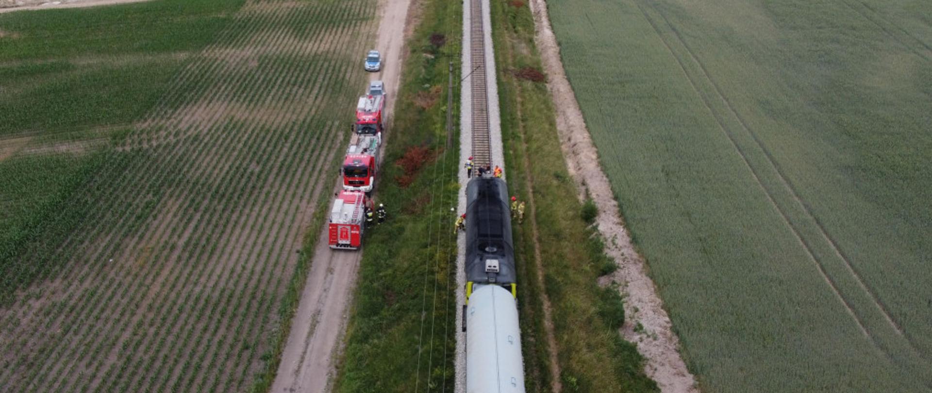 Zdjęcie przedstawia jednotorowy szlak kolejowy na której stoi lokomotywa z zespołem wagonów. Wokół lokomotywy widoczni strażacy, którzy wykonują zadania służbowe. Na drodze gruntowej równoległej do torowiska stoją samochody służb ratowniczych. 