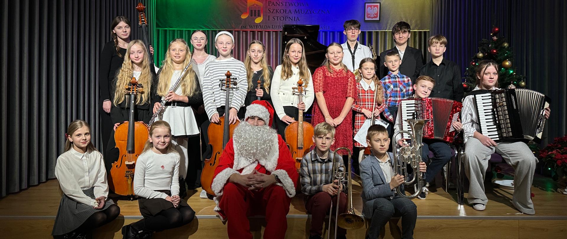 Na pierwszym planie siedzę dziewczynki i chłopcy. Chłopcy trzymają w rękach instrumenty puzon i sakshorn. Pomiędzy nimi siedzi Święty Mikołaj w czerwonym ubraniu z dużą białą brodą, a za nimi stoją dziewczynki i chłopcy w różnokolorowych ubraniach trzymają w rękach instrumenty wiolonczele, flet, saksofon, akordeon. W tle logo szkoły muzycznej.