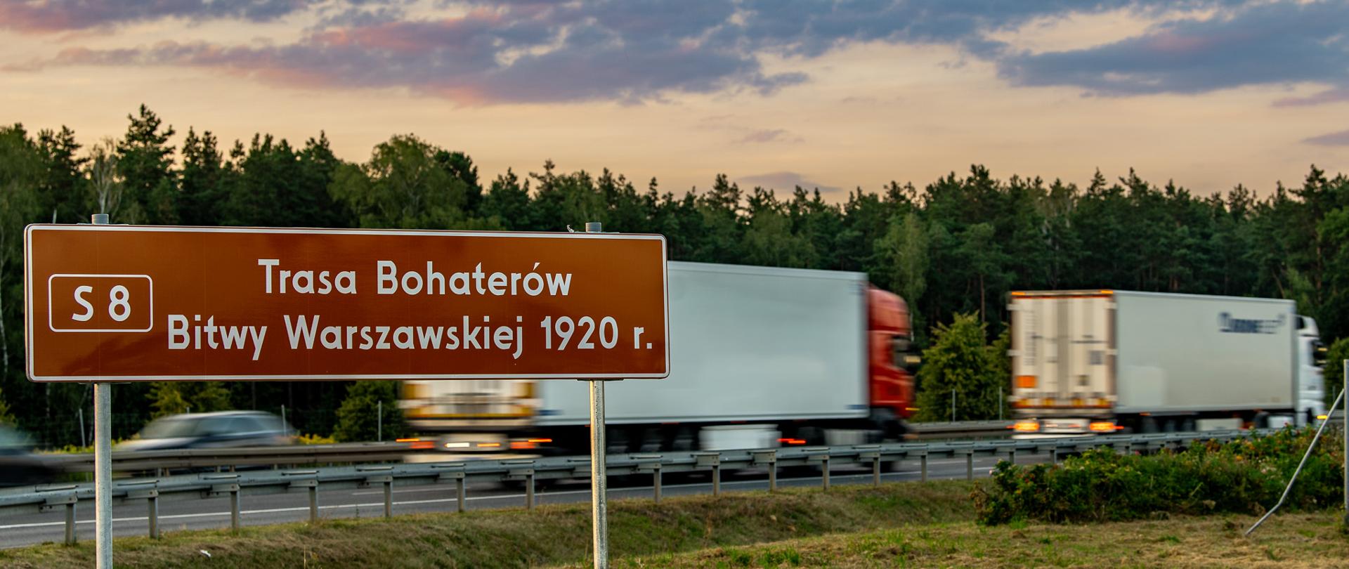 Zdjęcie przedstawiające tablicę informującą o poruszaniu się drogą ekspresową S8, Trasą Bohaterów Bitwy Warszawskiej 1920 r. W tle widać poruszające się drogą ekspresową pojazdy