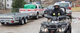 Strażak siedzący na pojeździe terenowym, w trakcie przygotowań do działań poszukiwawczych. W tle znajdują się samochody straży pożarnej. 