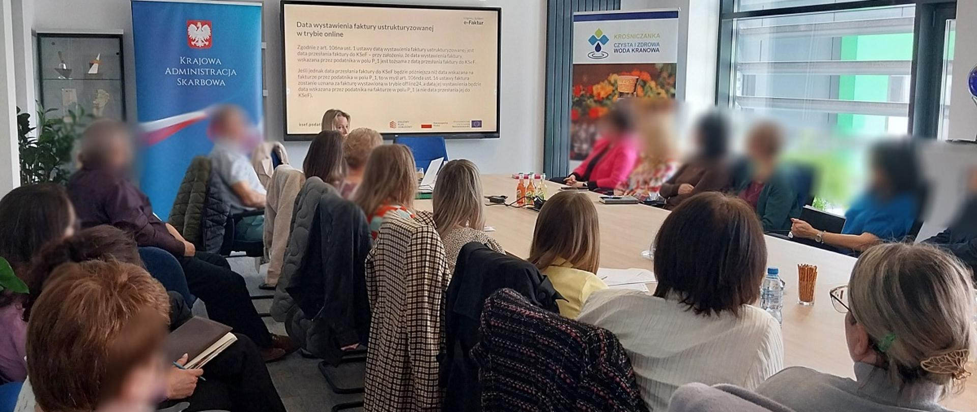 Sala konferencyjna. Przy stole siedzą uczestnicy szkolenia. W tle wyświetla się prezentacja. Po lewej stronie baner KAS.