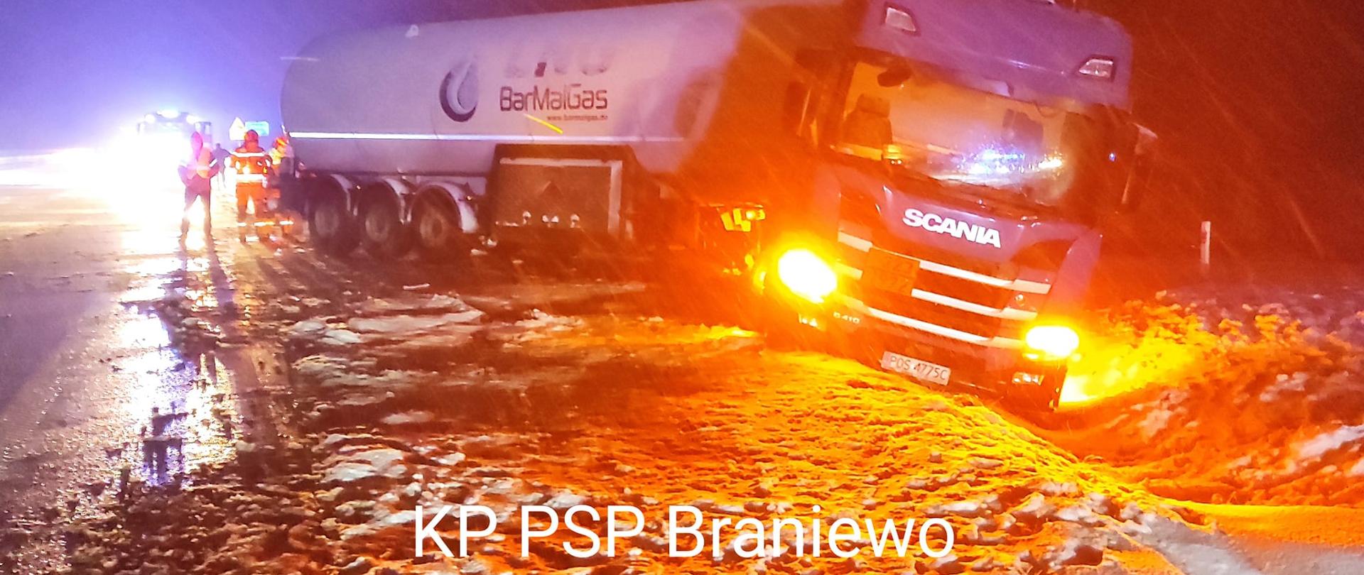 Zdjęcie zrobione nocą. Pada śnieg. Na zdjęciu auto cysterna stoi częściowo w rowie. Pobocze drogi w śniegu.