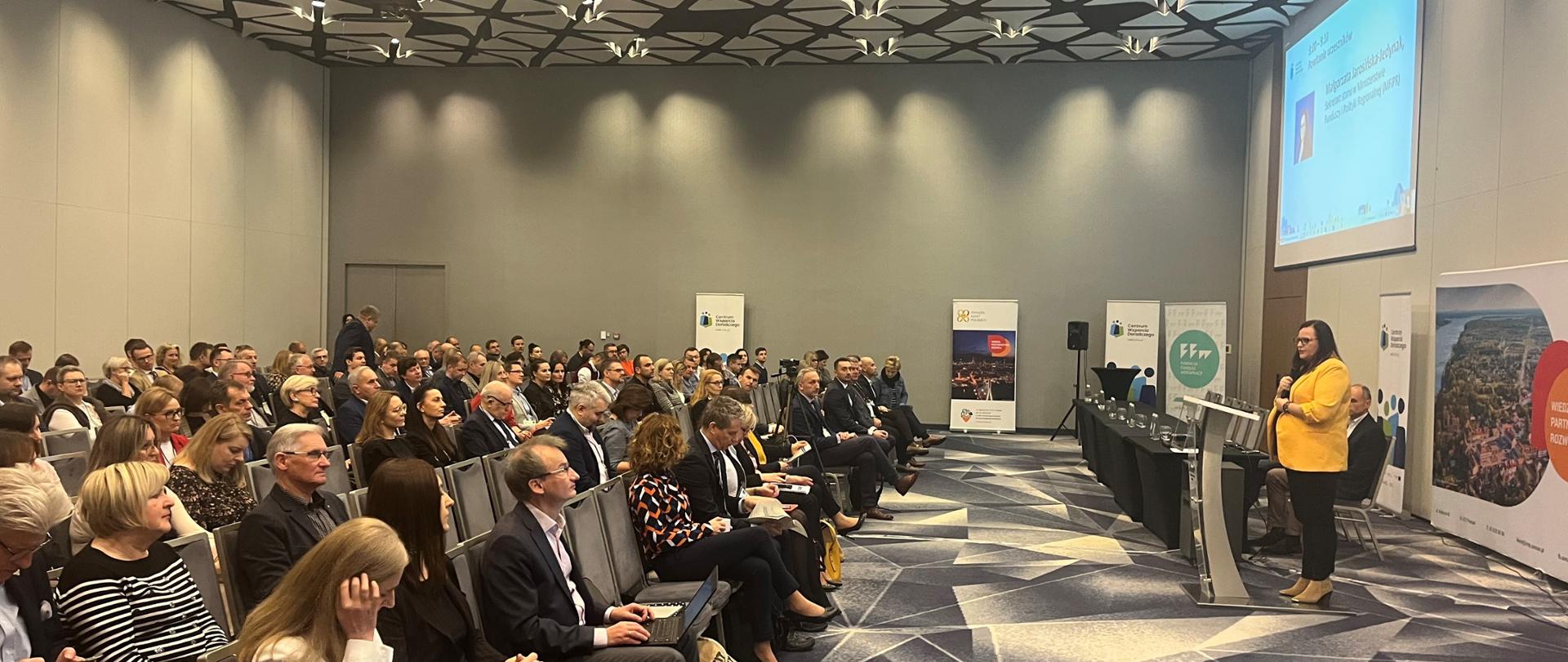 Na zdjęciu sala konferencyjna wypełniona siedzącymi słuchaczami. Po lewej przy mównicy stoi kobieta - wiceminister Małgorzata Jarosińska-Jedynak.