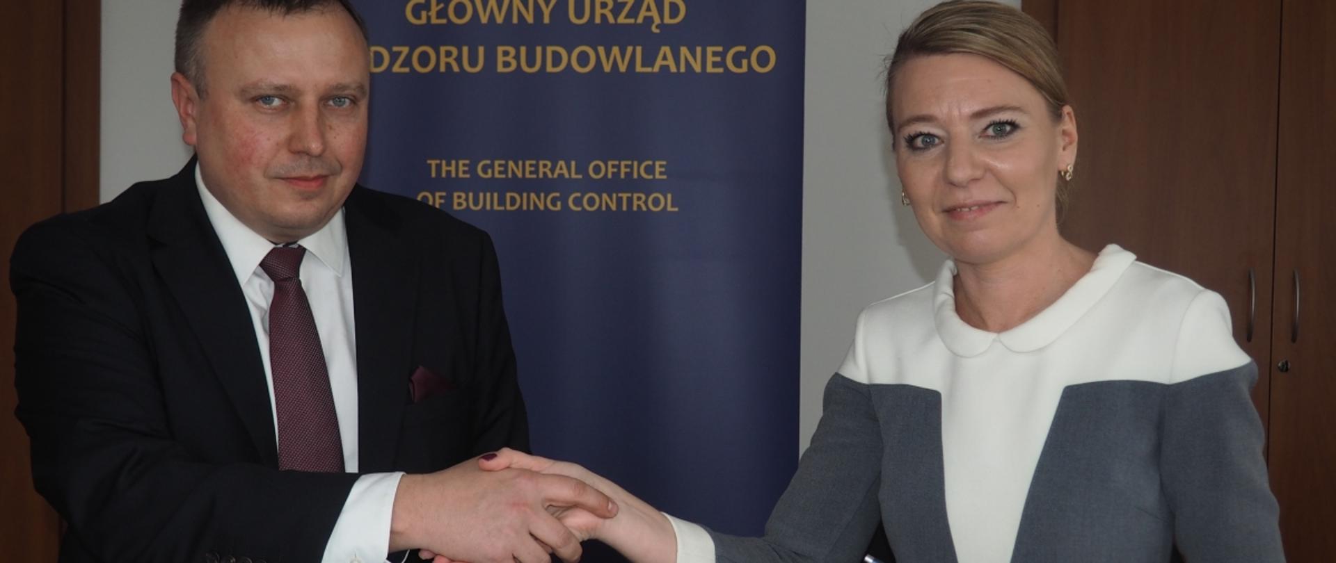 „Uścisk dłoni dwóch osób nad stołem z dokumentami, w tle baner GUNB.”