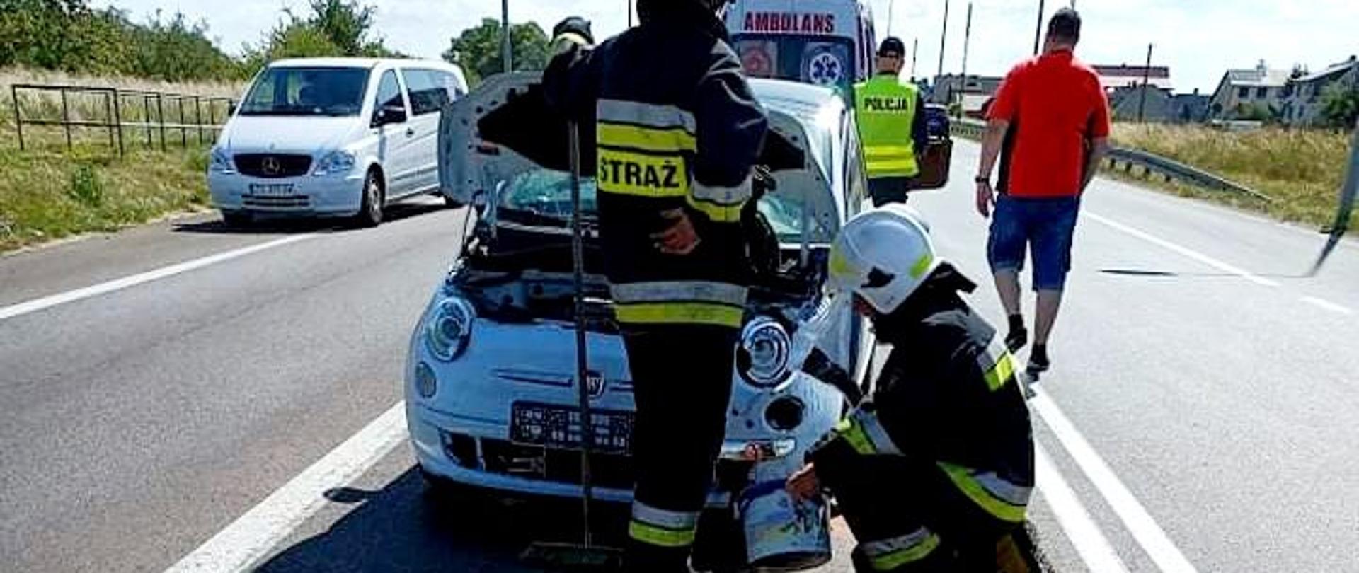 Zdjęcie przedstawia uszkodzony pojazd na drodze oraz 2 strażaków działających przy zdarzeniu. W tle widać karetkę.