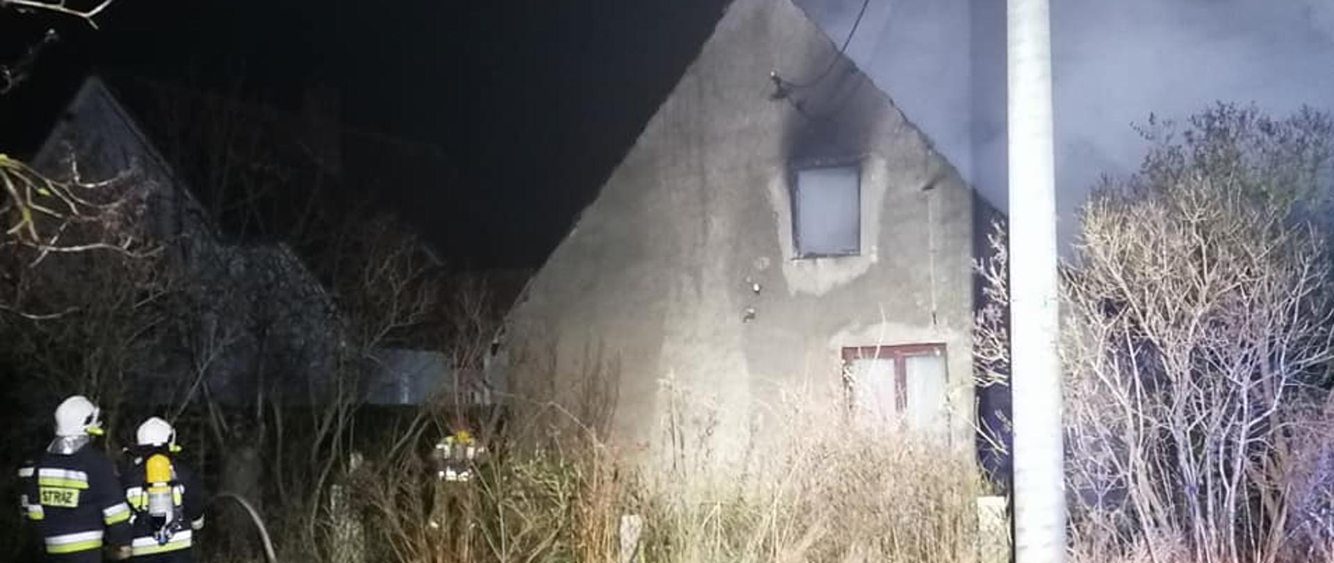 z lewej strony zdjęcia stoją dwaj strażacy ochotnicy zmierzający w stronę domu. Budynek widocznie zniszczony w oknie na piętrze widać całe pomieszczenie zadymione. Pora nocna, budynek oświetlony sztucznie.