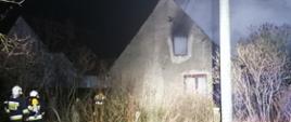 z lewej strony zdjęcia stoją dwaj strażacy ochotnicy zmierzający w stronę domu. Budynek widocznie zniszczony w oknie na piętrze widać całe pomieszczenie zadymione. Pora nocna, budynek oświetlony sztucznie.