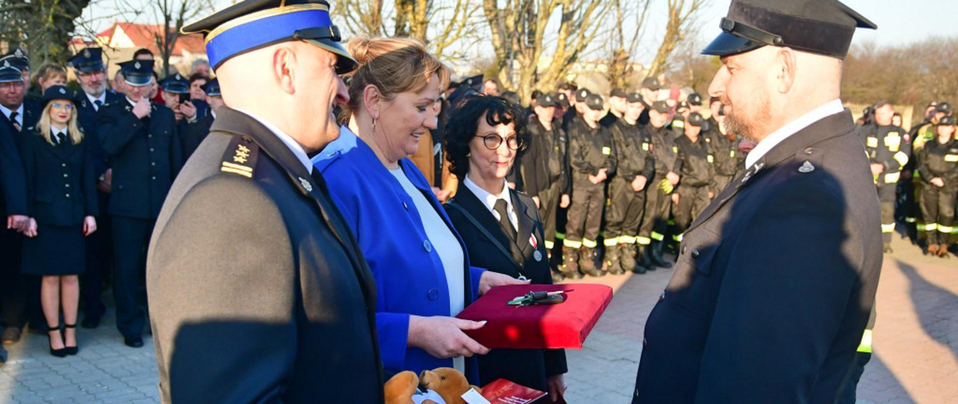 Oficer PSP - komendant, w ręku trzyma dokument i maskotkę. Z jego lewej strony dwie kobiety. Wszyscy składają gratulacje druhowi OSP. W tle kilkudziesięciu strażaków w mundurach galowych i koszarowych.