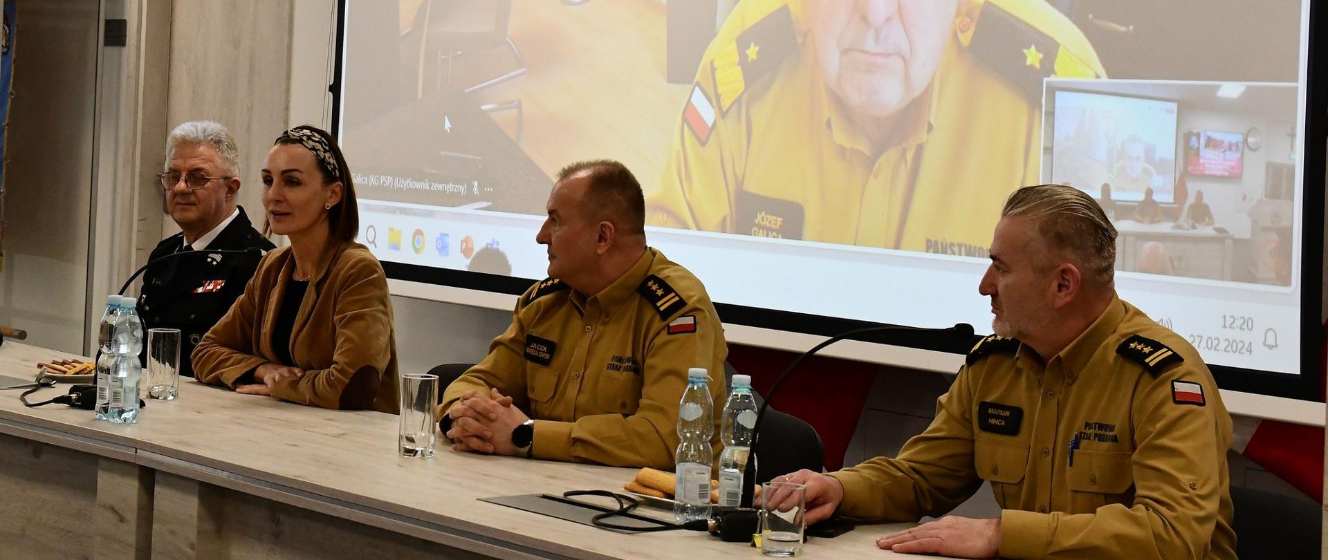 kobieta i trzech mężczyzn siedzą przy stole , w tle ekran a na nim mężczyzna w mundurze strażaka