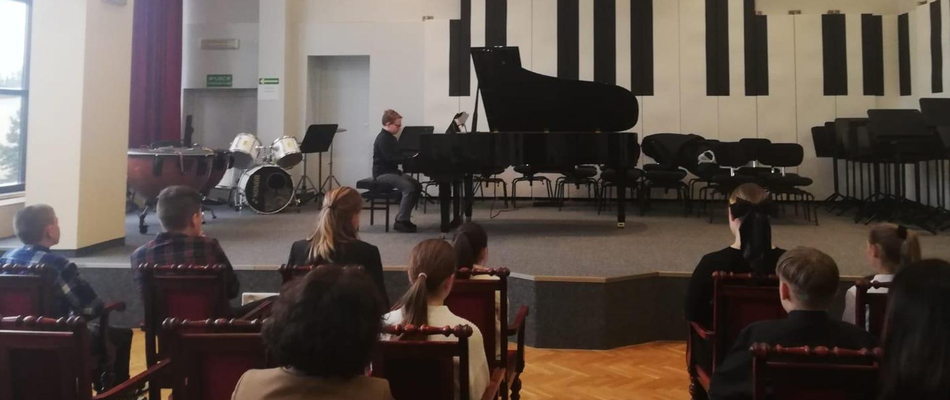 Chłopiec gra na fortepianie, z przodu widać publiczność.