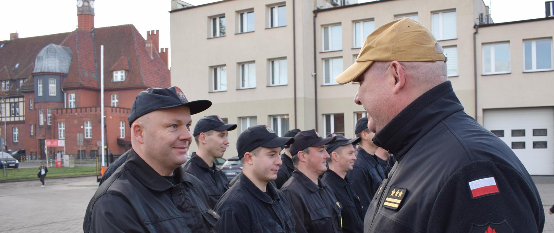 Komendant uściskuje dłoń strażaka stojącego w dwuszeregu. W tle budynek komendy i ratusz miasta.