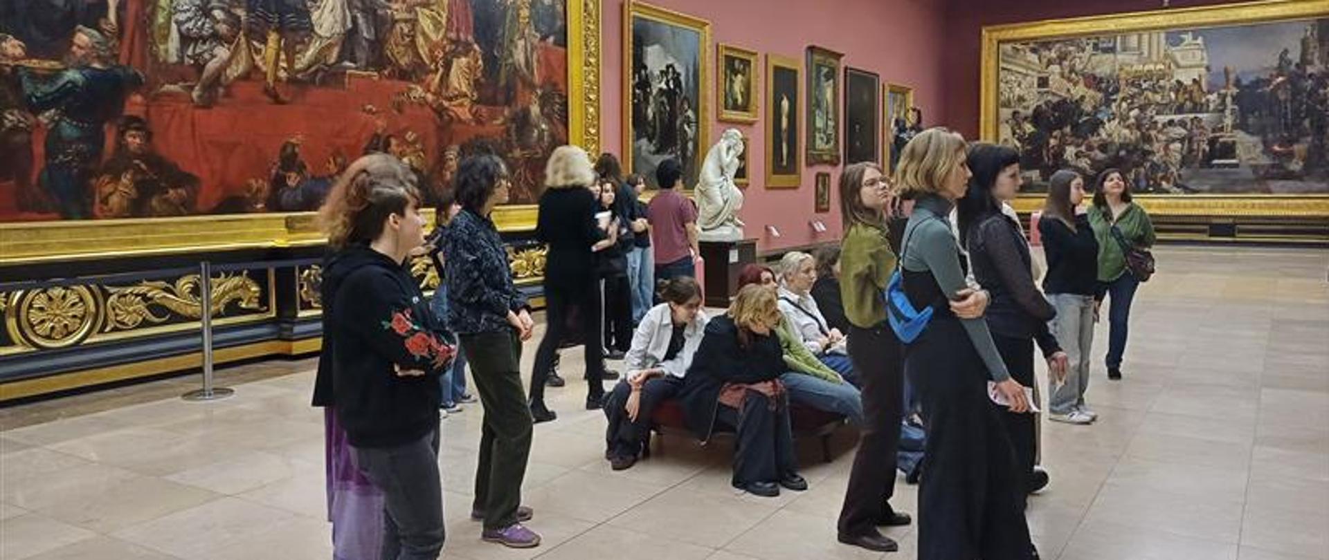 W dużej sali muzealnej na ścianach wiszą obrazy. W centrum sali zebrana jest grupa 20 młodych osób. Część stoi, a pozostali siedzą na niskiej kanapie, ustawionej na środku galerii. Oglądają eksponowane obrazy. Za grupą na ścianie wisi ogromnych rozmiarów „Hołd Pruski” Jana Matejki, a po prawej stronie, na krótszej ścianie, „Pochodnie Nerona” Henryka Siemiradzkiego. Pomiędzy tymi obrazami znajduje się kilka mniejszych dzieł. 