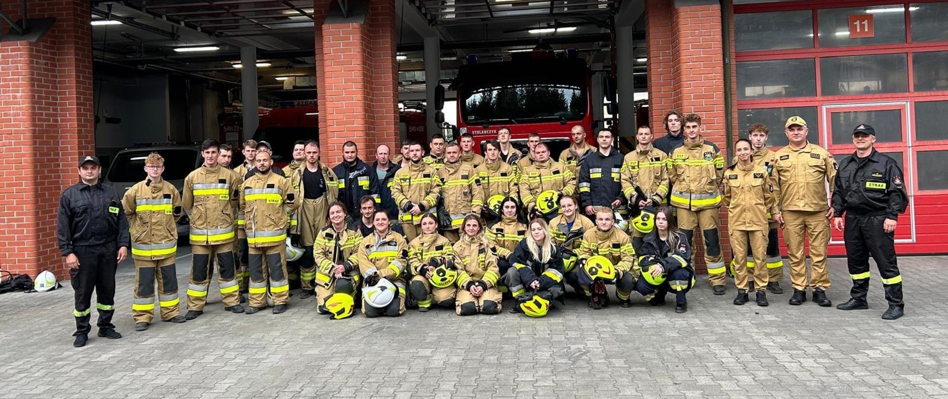Zdjęcie grupowe wykonane na zewnątrz. Przedstawia strażaków w umundurowaniu specjalnym. W tle garaż. 