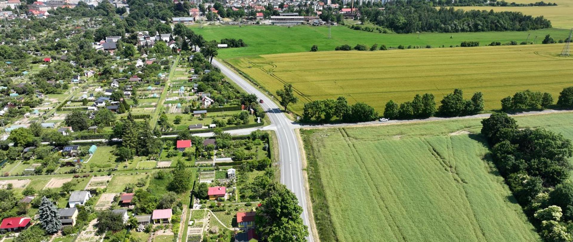Widok z lotu ptaka na wiejską okolicę. Po lewej stronie znajdują się ogródki działkowe z licznymi drzewami, krzewami i małymi domkami, w tym kilkoma z czerwonymi dachami. Po prawej stronie rozciągają się duże pola uprawne w różnych odcieniach zieleni i złota. Przez środek kadru biegnie asfaltowa droga, która przecina krajobraz i łączy obie części. W tle widać zabudowania miejskie oraz pas lasu.