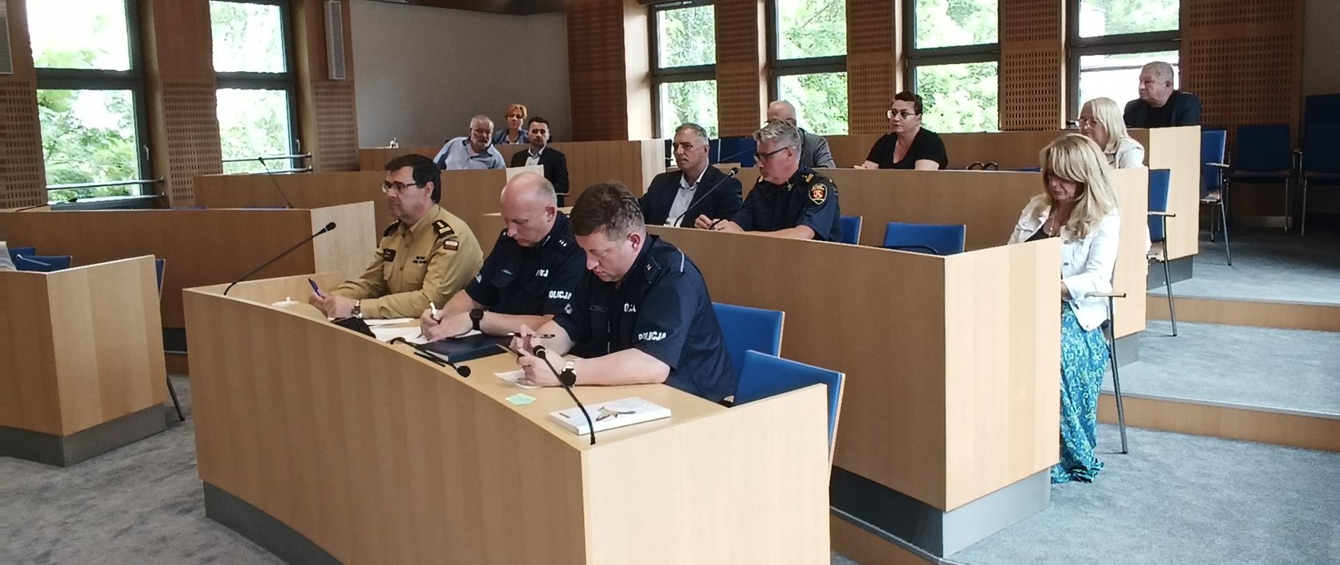 Sala sesyjna. Przy stołach widoczni przedstawiciele służb mundurowych oraz innych instytucji.