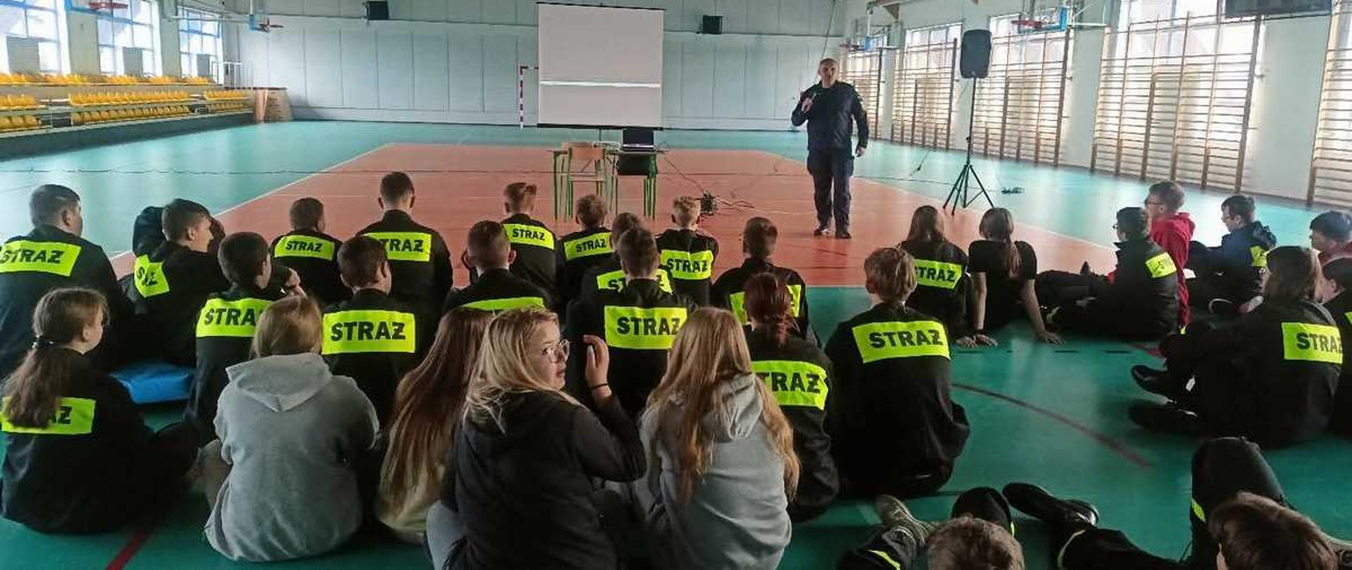 Zdjęcie przedstawia grupę młodzieży zgromadzoną w hali sportowej podczas spotkania informacyjnego lub szkolenia.
Kompozycja i sceneria
Pierwszy plan i środek: Widzimy plecy kilkudziesięciu młodych osób siedzących bezpośrednio na podłodze boiska. Większość z nich ma na sobie czarne bluzy lub mundury z jaskrawożółtymi, odblaskowymi pasami i dużym napisem „STRAŻ” na plecach. Sugeruje to, że są to członkowie Młodzieżowej Drużyny Pożarniczej.
Tło (centrum): Naprzeciwko grupy stoi mężczyzna w ciemnym mundurze, który przemawia do zgromadzonych. Obok niego znajduje się rozstawiony ekran projekcyjny oraz stolik z laptopem.
Otoczenie: Akcja dzieje się w dużej sali gimnastycznej. Po lewej stronie widać żółte trybuny, a po prawej drabinki gimnastyczne i duże okna, przez które wpada naturalne światło. Podłoga boiska jest zielono-pomarańczowa z wymalowanymi liniami do gier zespołowych.
Atmosfera
Zdjęcie oddaje nastrój skupienia i edukacji. Młodzież siedzi w luźnych grupach, niektórzy odwracają głowy, rozmawiając ze sobą, ale większość patrzy w stronę prelegenta. Całość ma charakter oficjalnego spotkania służb ratowniczych z młodymi adeptami.