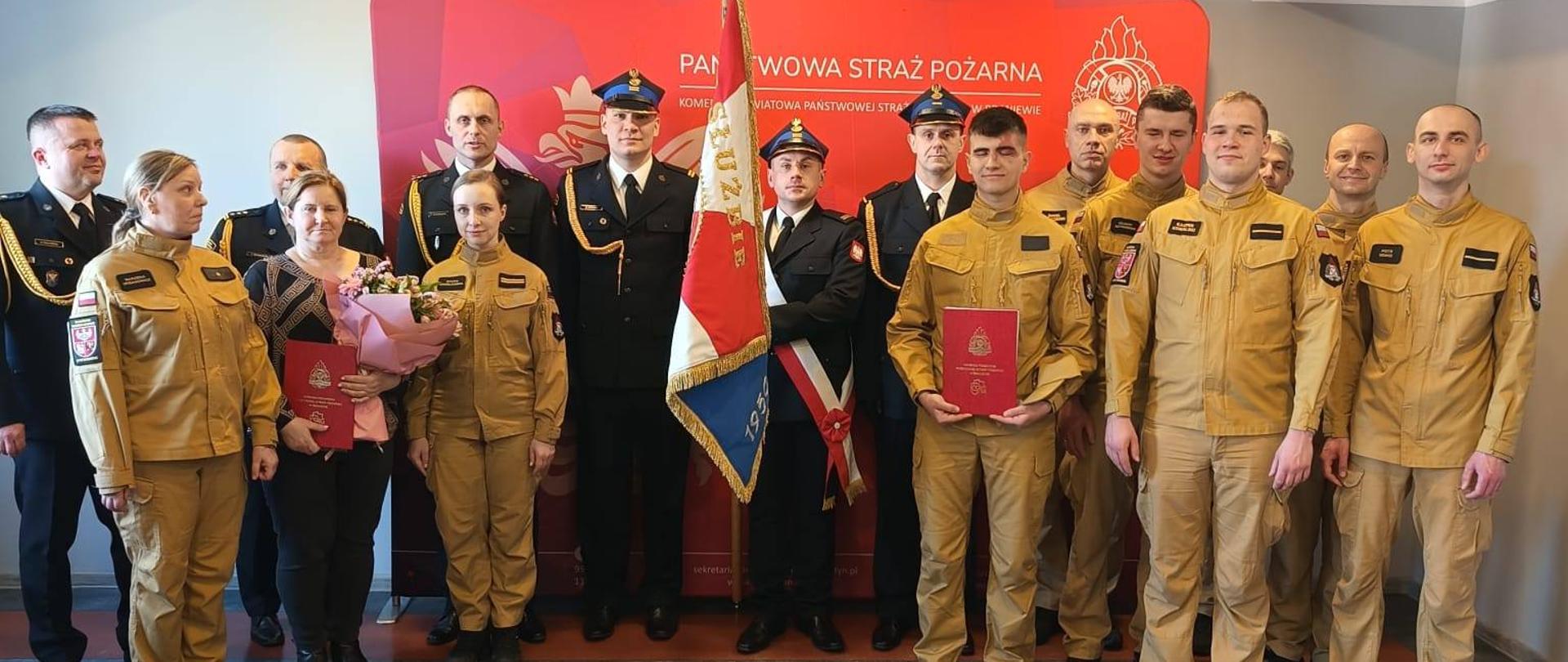 Grupa strażaków na świetlicy. Z prawej strony w jasnych mundurach i w galowych mundurach ze sztandarem.