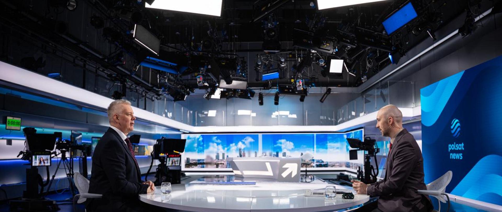 Na zdjęciu studio telewizyjne Polsat News. Na pierwszym planie dwie osoby siedzą przy białym, okrągłym stole - po lewej stronie minister Tomasz Siemoniak, a po prawej stronie - redaktor Marcin Fijołek. Na stole szklanki z wodą, gazety i inne przedmioty. U góry zdjęcia widać oświetlenia studia. Po prawej stronie ścianka multimedialna z wyświetlonym logo Polsat News (różne odcienie niebieskiego i granatowego). Wokół stołu kamery. W tle ścianka z widokiem miasta. 