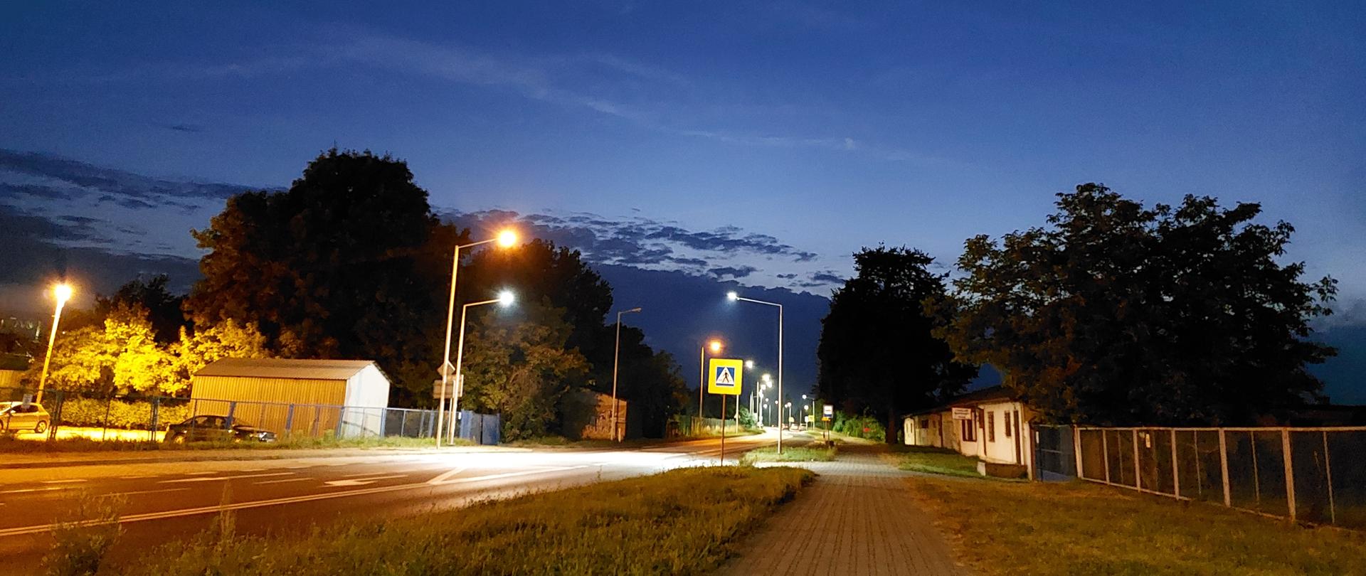 Przejście dla pieszych z nowym oświetleniem - droga w nocy wśród zabudowań, chodnik wzdłuż drogi, oznakowane i oświetlone przez latarnie przejście dla pieszych 