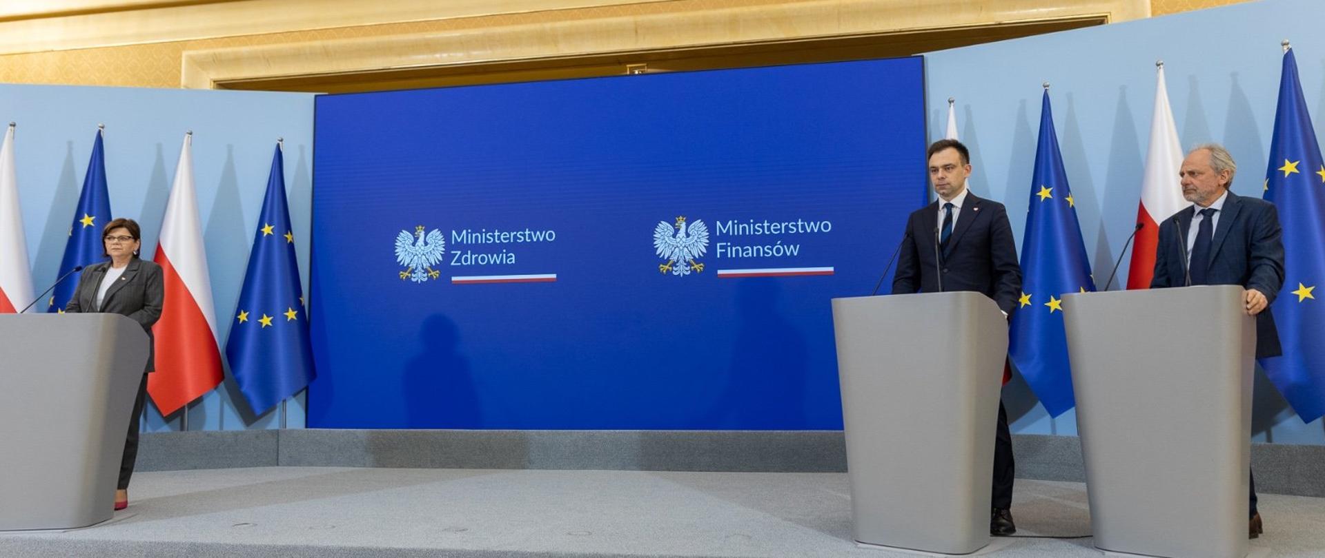 Minister finansów Andrzej Domański, minister zdrowia Izabela Leszczyna,
wiceminister finansów Jarosław Neneman stoją za mównicami, w tle flagi Polski i UE