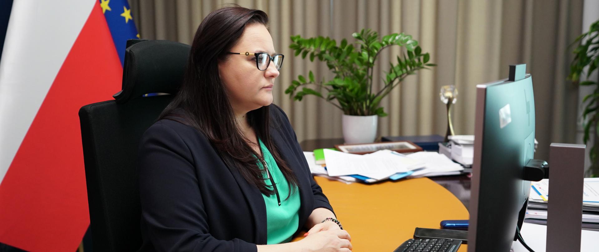 Wiceminister Małgorzata Jarosińska-Jedynak w gabinecie siedzi przed monitorem. Za nią flagi PL i UE. Obok kwiaty w doniczce.