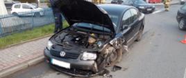 Zdjęcie przedstawia uszkodzony samochód osobowy marki volkswagen. Na zdjęciu widać uszkodzony prawy przód samochodu, rozbity prawy reflektor, uszkodzony zderzak przedni oraz zniszczony prawy błotnik wraz z nadkolem i kołem. Podniesiona jest także maska samochodu. Samochód znajduje się przy krawężniku po prawej stronie drogi. Za samochodem po lewej stronie widać policjanta zbierającego informacje z osobą kierującą obok nich stoi strażak. Po prawej stronie widać przejeżdżające samochody oraz strażaka kierującego ruchem.