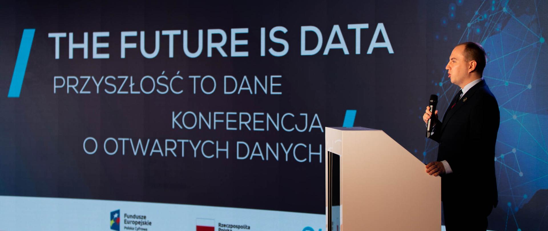 Wiceminister Adam Andruszkiewicz przemawia z pulpitu. W tle napis: The future is data. Przyszłość to dane. Konferencja o otwartych danych.