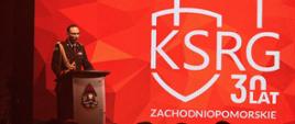 Strażak w galowym mundurze stoi za mównicą na tle dużego czerwonego ekranu z napisem KSRG 30 lat Zachodniopomorskie. Przed sceną siedzi publiczność