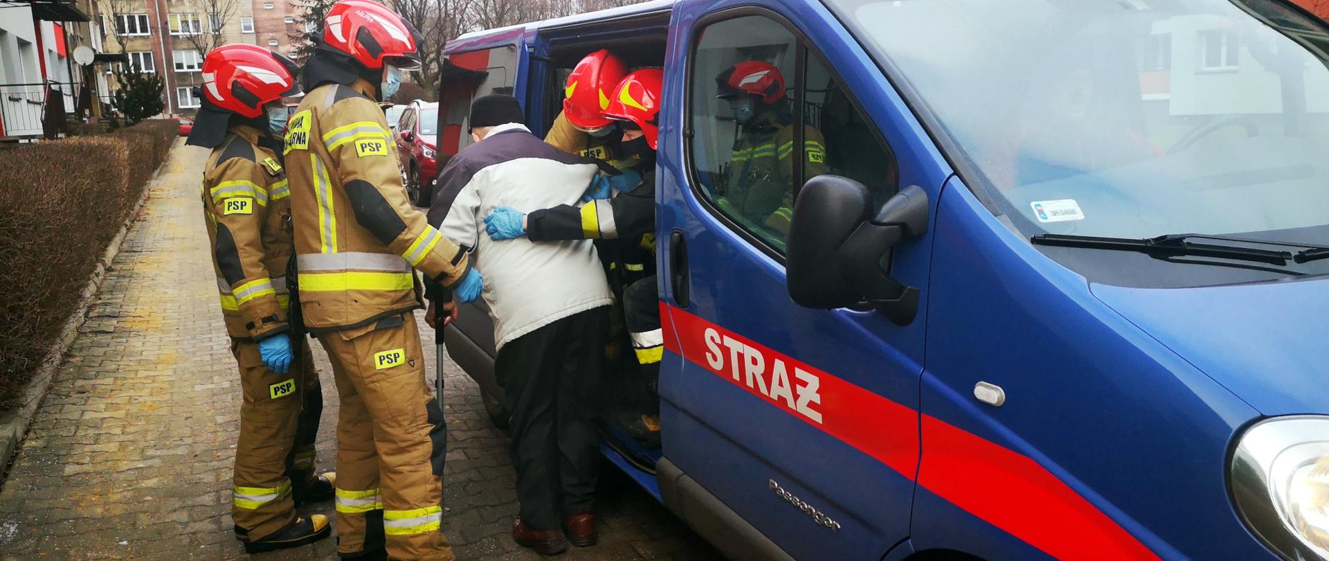 Zdjęcie przedstawia strażaków pomagających starszemu panu w dotarciu do punktu szczepień. czterech strażaków ubezpiecza pana podczas wysiadania z pojazdu.